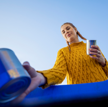 Vista de dentro de uma lixeira de reciclagem, uma jovem mulher com tranças deposita latas de alumínio.